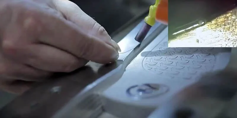 Primer plano de una mano utilizando una herramienta para realizar grabados intrincados sobre metal, demostrando una precisión similar a la de la soldadura láser en el sector de los moldes. Un pequeño recuadro revela una vista detallada del proceso de grabado.