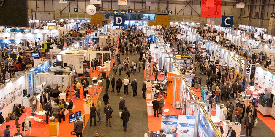 Una feria comercial grande y animada con numerosos stands y asistentes caminando por los pasillos de una sala de exposiciones.