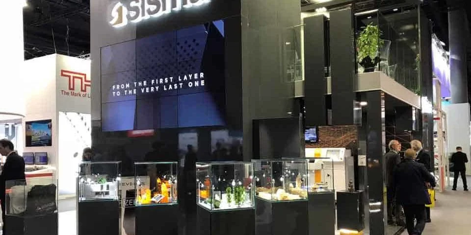 Stand de feria con vitrinas y una gran pantalla con el texto: "De la primera capa a la última". Varias personas observan las exhibiciones que destacan la fabricación aditiva.