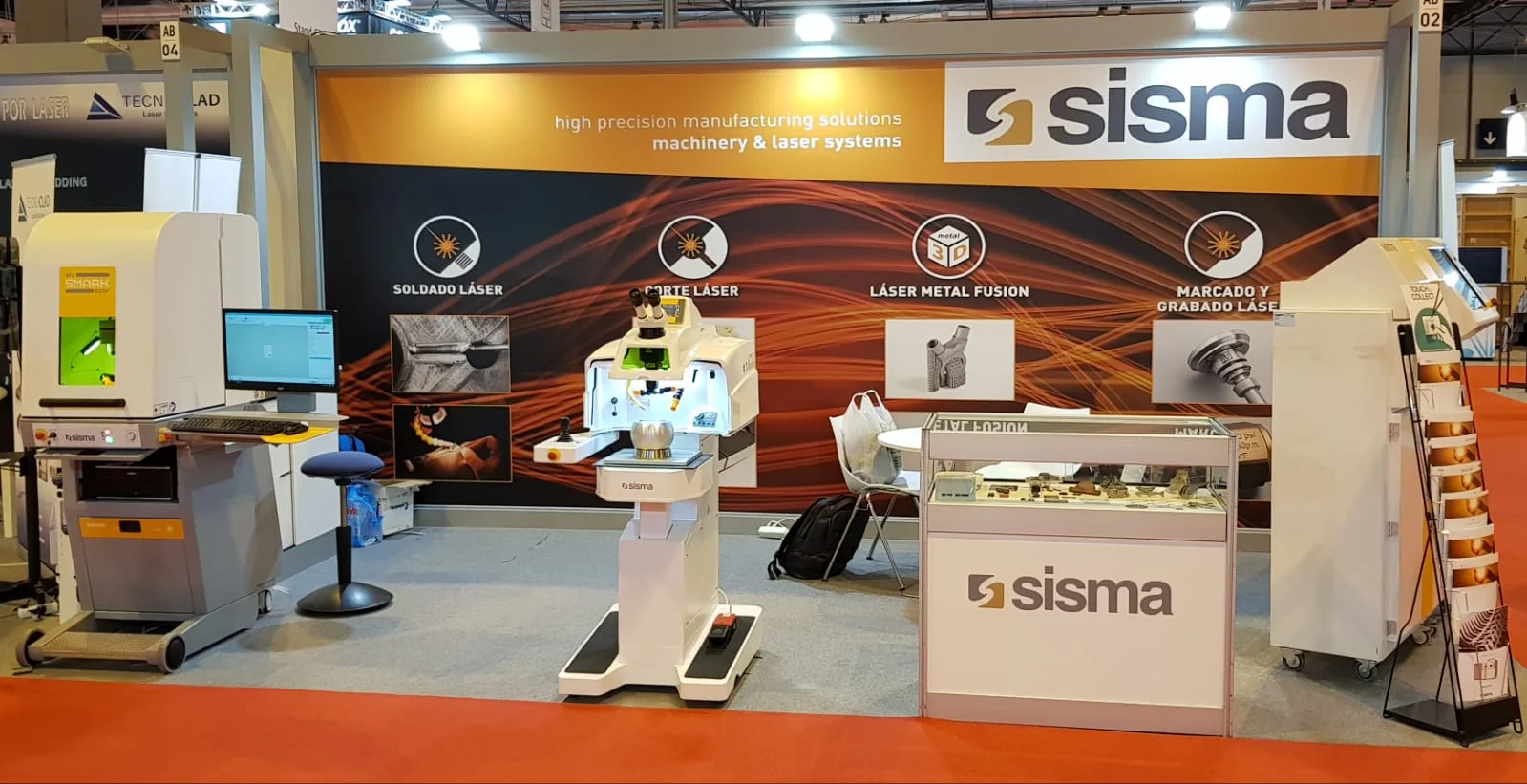 Stand de exposición con maquinaria láser, displays y folletos de Sisma sobre una mesa, con un fondo que resalta las soluciones de fabricación.