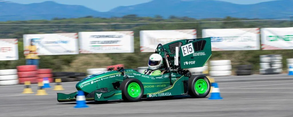 Un coche de carreras verde con la marca "UPC Terrassa" maniobra hábilmente alrededor de conos azules y amarillos en la pista, mostrando una precisión similar a la soldadura láser en la automoción, todo ello frente a un impresionante paisaje montañoso.