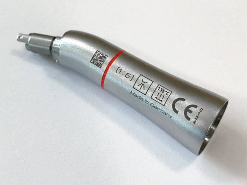 Una pieza de mano dental plateada con un código QR, símbolos y el texto "Made in Germany" impresos en su lateral, colocada sobre una superficie blanca.