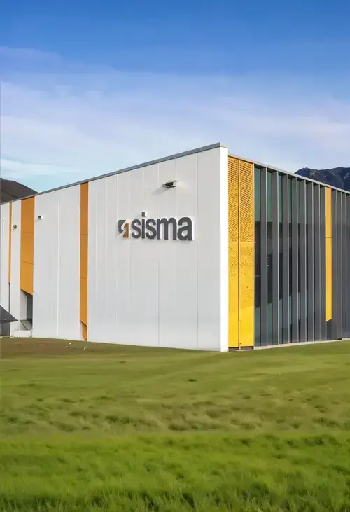 Moderno edificio industrial con grandes ventanales y detalles en naranja, con el logotipo "sisma" en la pared exterior. Se ve hierba verde y cielo azul despejado.