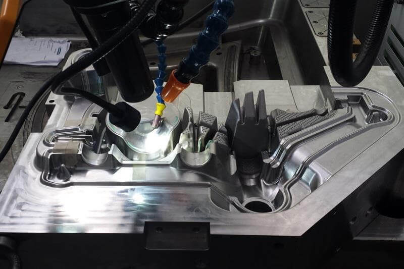 Vista en primer plano de una máquina herramienta CNC que fresa una detallada pieza de molde metálica reflectante en un entorno industrial.