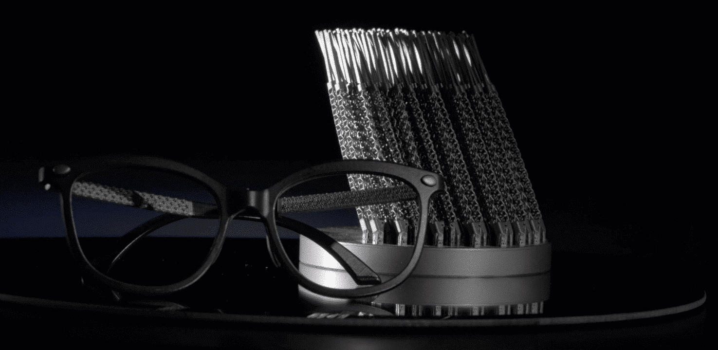 Un par de gafas negras con patillas texturizadas se exhiben delante de un soporte cilíndrico que sostiene varias patillas idénticas, sobre un fondo oscuro.