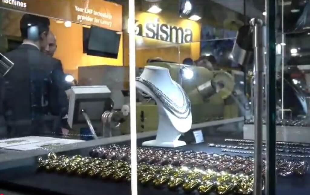 Vitrina de joyería con collares y pulseras, con personas y equipos visibles al fondo en la 31ª BIEMH 2022.