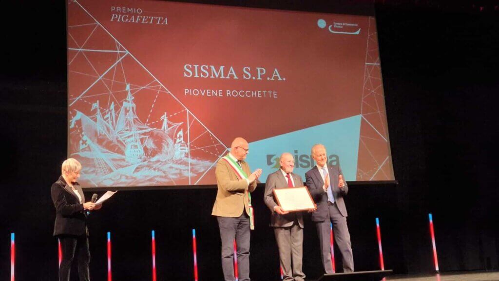 Cuatro personas de pie en el escenario durante una ceremonia de entrega de premios. Un hombre sostiene un certificado enmarcado delante de una pantalla que muestra "SISMA S.P.A. PIOVENE ROCCHETTE", reconocido por la innovación en soldadura láser en la automoción.