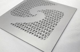 La plantilla de metal con numerosos patrones circulares pequeños crea una gran letra "S" estilizada sobre una superficie lisa y gris claro.