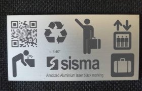 Letrero metálico con iconos: código QR, símbolo de reciclaje, persona inclinada sobre lavabo, persona con equipaje, ascensor, maleta y texto "sisma". Marcado láser negro en aluminio anodizado.