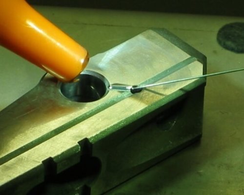 Primer plano de una pistola de calor que se utiliza para aplicar calor a un componente metálico sujetado en un bloque metálico de tornillo de banco, con un alambre colocado cerca.