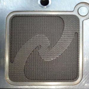 Un primer plano de una superficie metálica texturizada muestra un patrón abstracto y arremolinado grabado en forma cuadrada. El intrincado diseño, que evoca el marcado y grabado en moldes y matrices, está elegantemente rodeado por un borde plateado con una pestaña circular en la parte superior derecha.