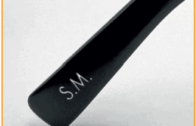 Primer plano de una patilla de gafas de color negro con la palabra "S.M." grabada sobre un fondo blanco liso.
