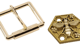 Hebilla de cinturón rectangular dorada junto a un medallón hexagonal con diseño de abeja.