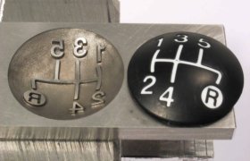 Dos piezas de patrón de cambio de marchas: una de metal con símbolos en relieve, una de plástico negro con símbolos blancos, que muestran diseños de transmisión manual con una "R" para reversa.