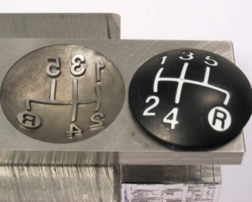 Dos piezas de patrón de cambio de marchas: una de metal con símbolos en relieve, una de plástico negro con símbolos blancos, que muestran diseños de transmisión manual con una "R" para reversa.