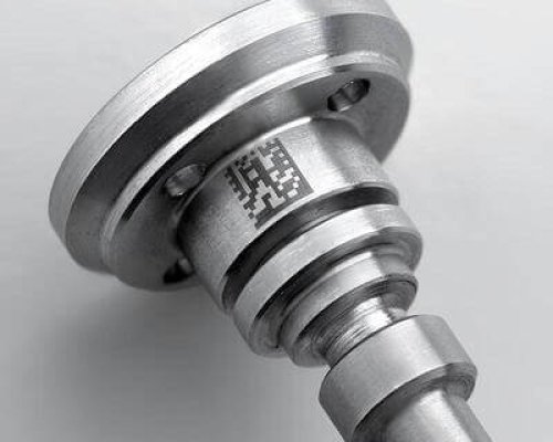Primer plano de una pieza industrial metálica con un código matricial único en blanco y negro grabado en su superficie cilíndrica.