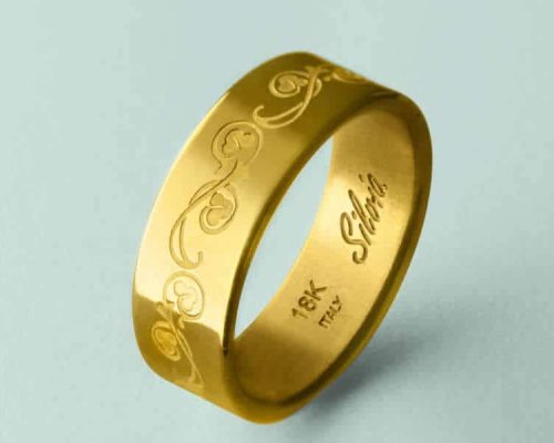 Anillo de oro con diseño de espiral grabado, con la inscripción "18K Italia" y "Silvia". Fondo azul pálido.