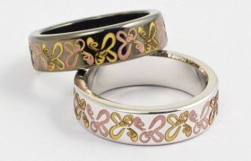 Dos anillos entrelazados con diseños de mariposas en rosa y dorado sobre bandas metálicas sobre un fondo blanco liso.