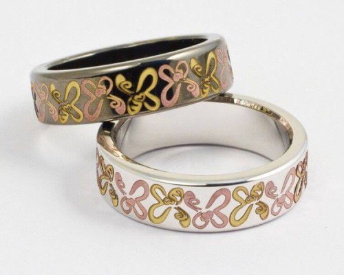 Dos anillos entrelazados con diseños de mariposas en rosa y dorado sobre bandas metálicas sobre un fondo blanco liso.