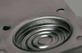 Primer plano de un componente metálico con ranuras circulares y dos orificios en una brida cuadrada, probablemente parte de una máquina o dispositivo electrónico.