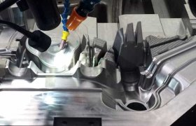 Primer plano de una máquina herramienta CNC cortando una pieza metálica, donde se aprecian ranuras y patrones intrincados en la superficie metálica. Se ven varios cabezales y mangueras alrededor de la pieza.