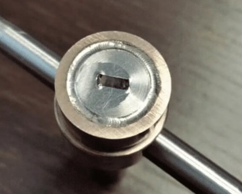 Primer plano de una cerradura metálica circular con bocallave rectangular, sujeta a una barra metálica, colocada sobre una superficie de madera.