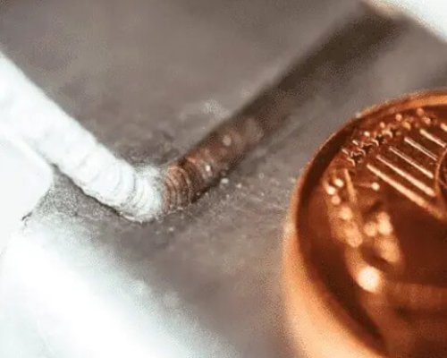 Primer plano de una pequeña soldadura metálica en una superficie metálica junto a una moneda de cobre, que muestra el detalle y la escala de la soldadura.