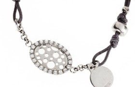 Una pulsera de plata que presenta un colgante ovalado con recortes circulares y pequeñas piedras preciosas, unido a un cordón negro con una etiqueta plateada redonda.