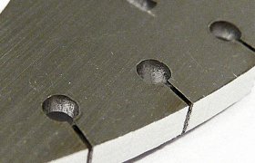 Primer plano de un objeto metálico con tres agujeros circulares y ranuras paralelas, sobre un fondo blanco.