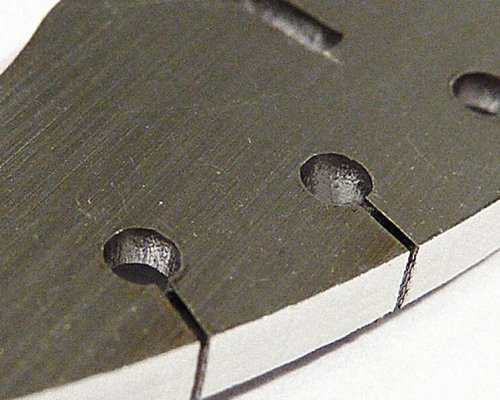 Primer plano de un componente metálico con orificios taladrados y ranuras rectas, que muestra una superficie y un borde mecanizados sobre fondo blanco.