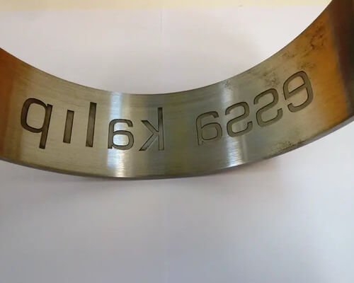 Superficie metálica curvada con el texto grabado "pllik a 955" representado al revés, colocado sobre un fondo blanco liso.