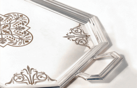 Una bandeja de plata que presenta un diseño floral adornado en el centro y patrones geométricos decorativos en las esquinas, con un asa de forma única.