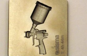 Placa metálica grabada que representa una pistola pulverizadora con un bote de pintura. La palabra "asma" y la hora "T: 4h 44m" están marcadas en la superficie de la placa.