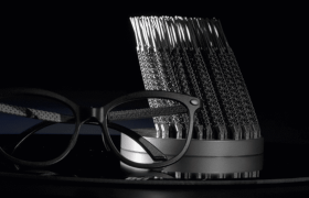 Unas gafas negras y una estructura cilíndrica de metal con superficie texturizada se colocan sobre una superficie negra reflectante, sobre un fondo oscuro.