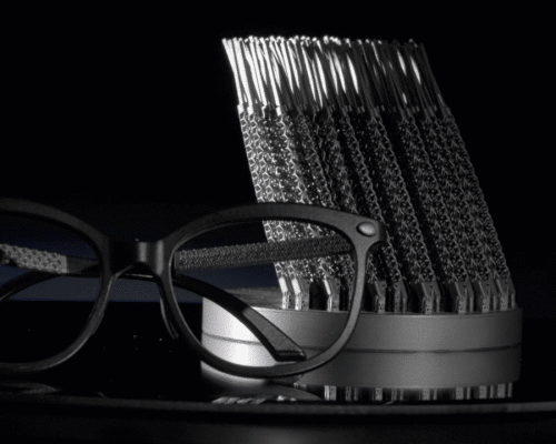 Un par de gafas negras con patillas texturizadas se exhiben delante de un soporte cilíndrico que sostiene varias patillas idénticas, sobre un fondo oscuro.