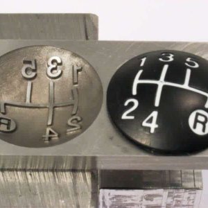 Dos placas redondas con patrón de cambio de marchas: la izquierda es metálica con números en relieve y la derecha es negra con números blancos impresos. Ambas muestran un patrón de 5 velocidades con reversa.