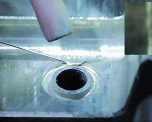 Primer plano de una soldadura TIG en curso sobre una superficie metálica cerca de una abertura circular, con un recuadro que muestra una vista ampliada de la unión soldada.