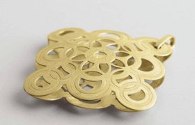 Un colgante de metal de color dorado con un intrincado diseño de patrón circular sobre un fondo gris claro.