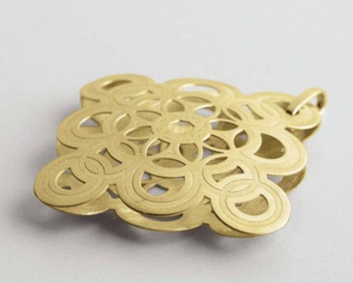 Un colgante de metal de color dorado con un intrincado diseño de patrón circular sobre un fondo gris claro.